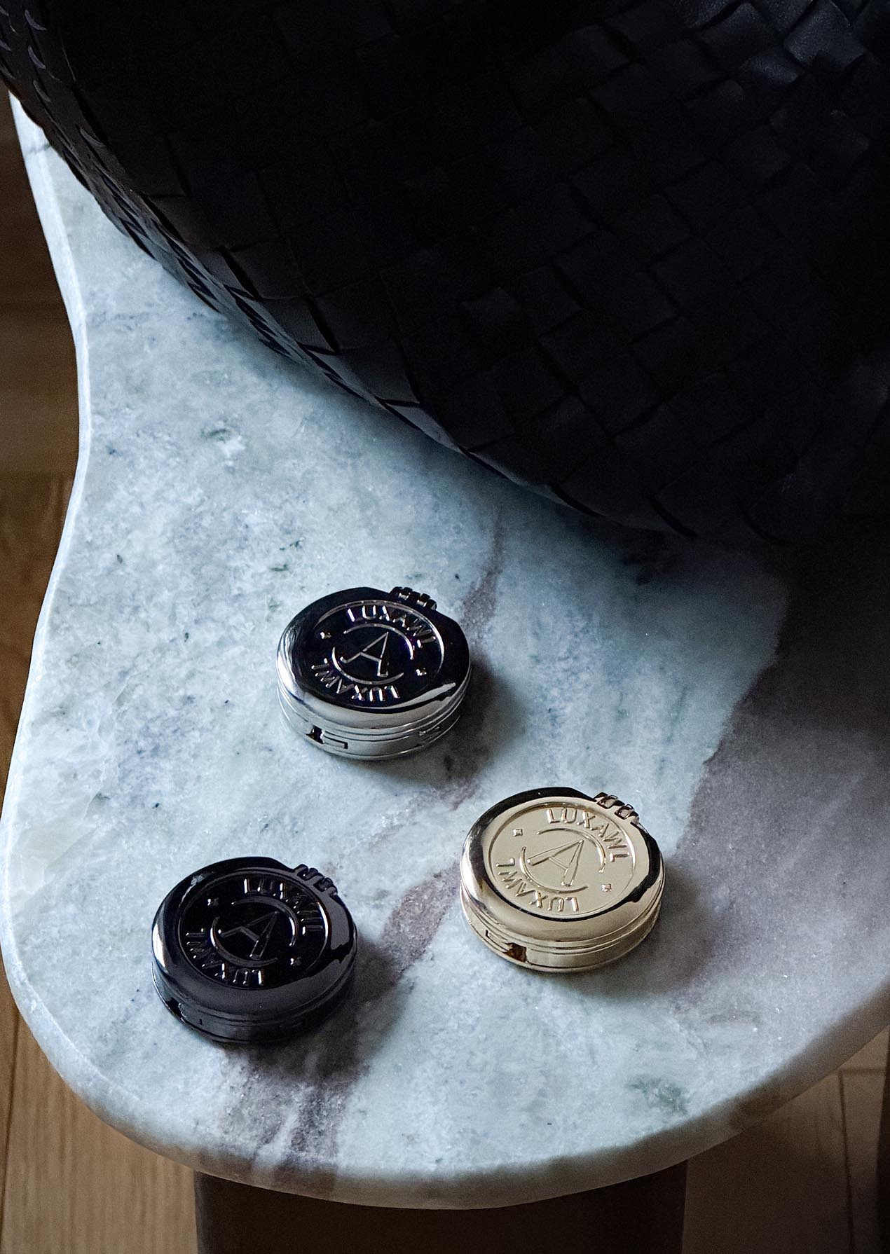 Gold silver and black LUXAWL bag hooks on a white marble table next to a black leather woven bag